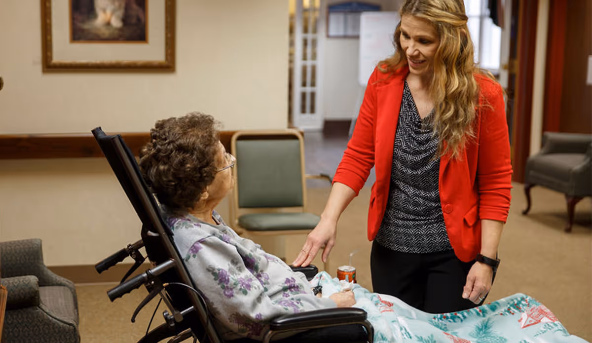 A woman in a red cardigan is smiling and gently touching the hand of an elderly woman sitting in a wheelchair, covered with a floral blanket, inside a senior living facility room with chairs and a painting on the wall.