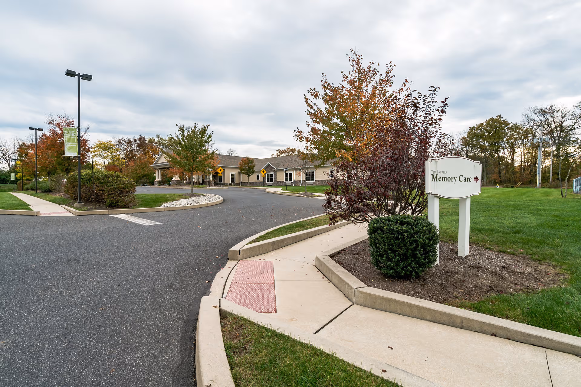 Curved driveway and sidewalk leading to a single-story building surrounded by trees with autumn foliage. A sign near the sidewalk reads 'The Laurels Memory Care' with an arrow pointing to the right. The sky is cloudy.