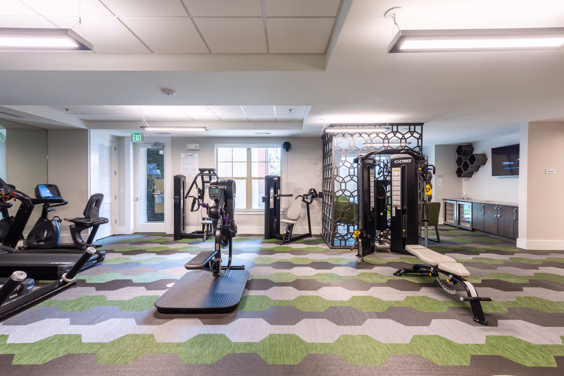 A well-lit fitness room with various exercise equipment including treadmills, stationary bikes, weight machines, and benches. The floor has a patterned carpet in shades of green, gray, and brown. There is a decorative partition and a wall-mounted TV in the background.