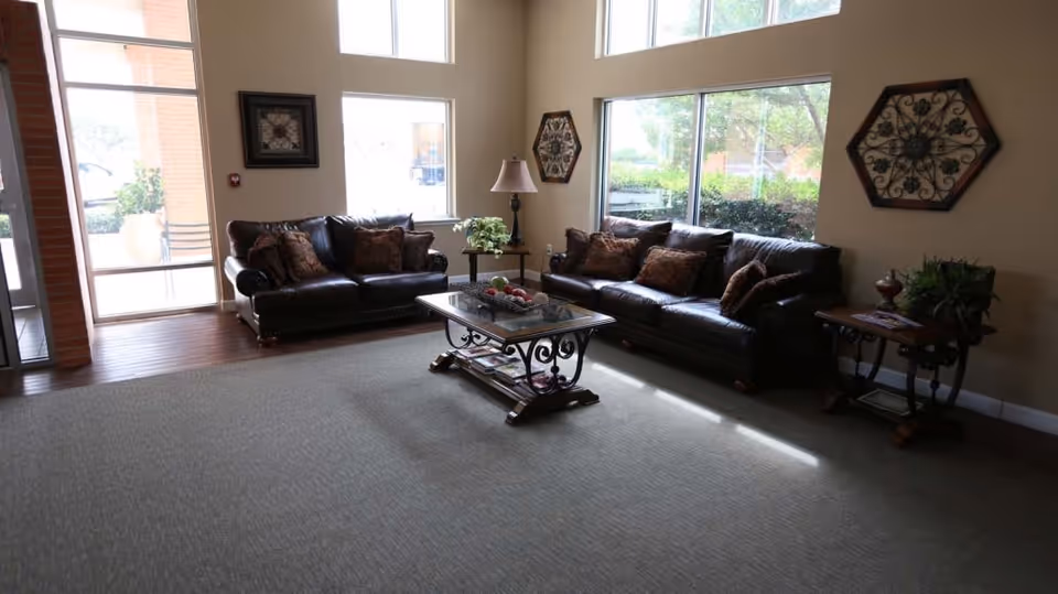 A cozy living room area with two dark leather sofas adorned with decorative pillows, a glass-top coffee table with ornamental legs, and two side tables with plants and a lamp. Large windows allow natural light to fill the room, and decorative wall art hangs on the beige walls.