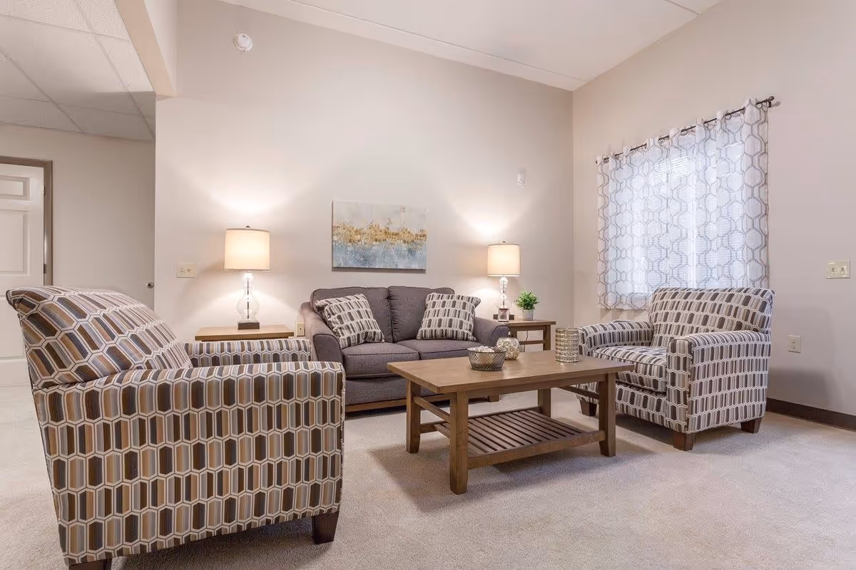 A cozy living room with a gray sofa and two patterned armchairs arranged around a wooden coffee table. Two table lamps with beige shades sit on side tables beside the sofa, and a window with patterned curtains lets in natural light. A piece of abstract artwork hangs on the wall above the sofa.