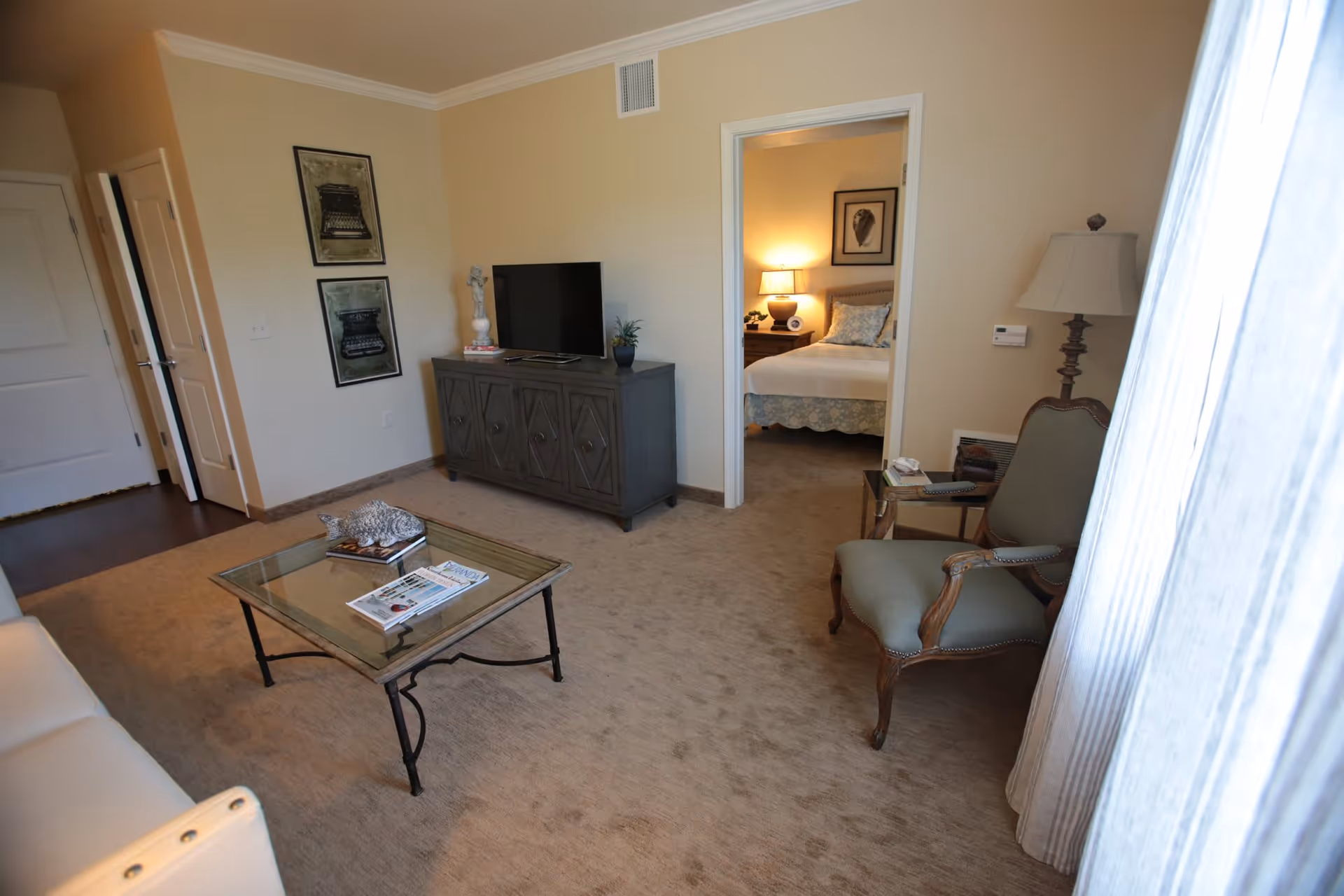 A cozy living room with beige carpet and walls, featuring a glass coffee table with magazines and a decorative item, a gray TV stand with a flat-screen TV and decorative pieces, a green upholstered armchair with wooden frame, a floor lamp, and a view into a bedroom with a bed, nightstand, lamp, and framed artwork.