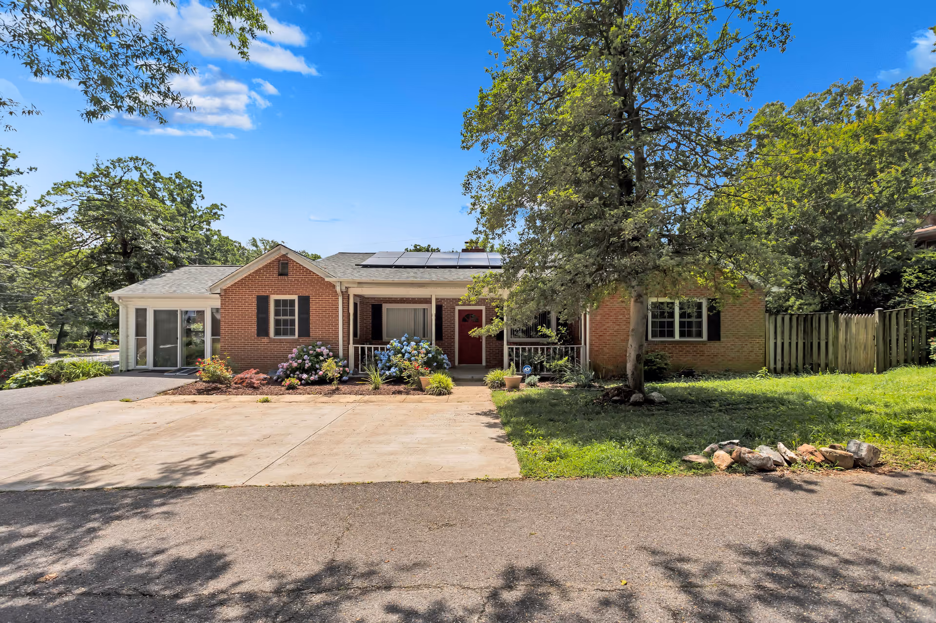 Single-story brick house front with a driveway, small porch, flowering shrubs, a tree, and solar panels on the roof under a blue sky.