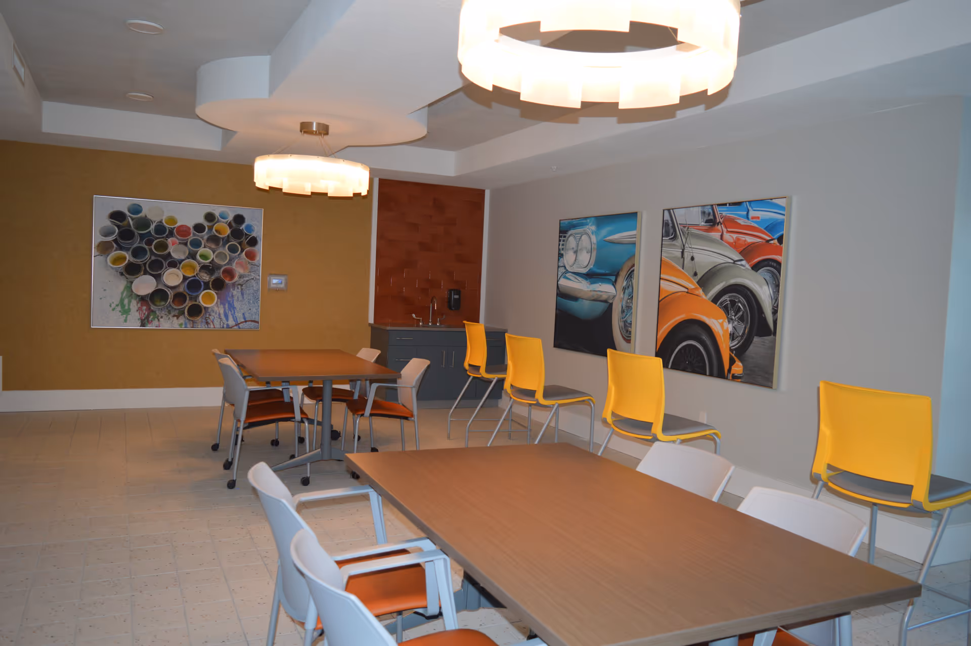 A modern dining area with two rectangular wooden tables surrounded by chairs, some with orange seats and others with yellow backs. The room features two circular ceiling lights, a small sink area with a red tiled backsplash, and colorful artwork on the walls including a heart made of paint cans and paintings of vintage cars.