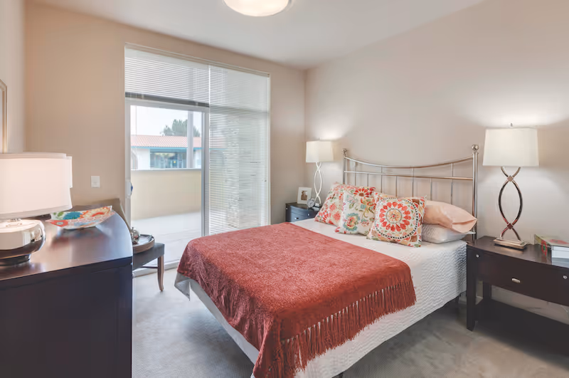 A bright and cozy bedroom with a bed covered in a white quilt and a red throw blanket. The bed has three decorative pillows with colorful patterns. There are two dark wooden nightstands on either side of the bed, each with a modern lamp. A dresser with a lamp and decorative items is on the left side of the room. A large window with blinds lets in natural light and opens to a small balcony with a view of neighboring buildings.