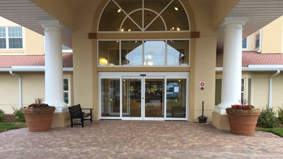 Entrance of a building with large white columns on either side, two large flower pots with red flowers, a black bench, and automatic sliding glass doors under an arched window.