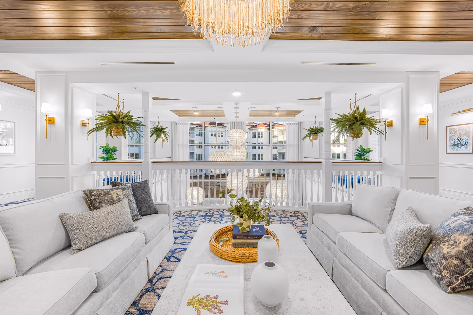 A bright and elegant living room area with two light gray sofas facing each other, adorned with decorative pillows. A white coffee table between the sofas holds a wicker tray, books, a plant, and white vases. The room features white walls with paneling, hanging green plants, wall sconces with warm lighting, and a wooden ceiling with a large chandelier. Large windows in the background reveal an exterior view of a building.