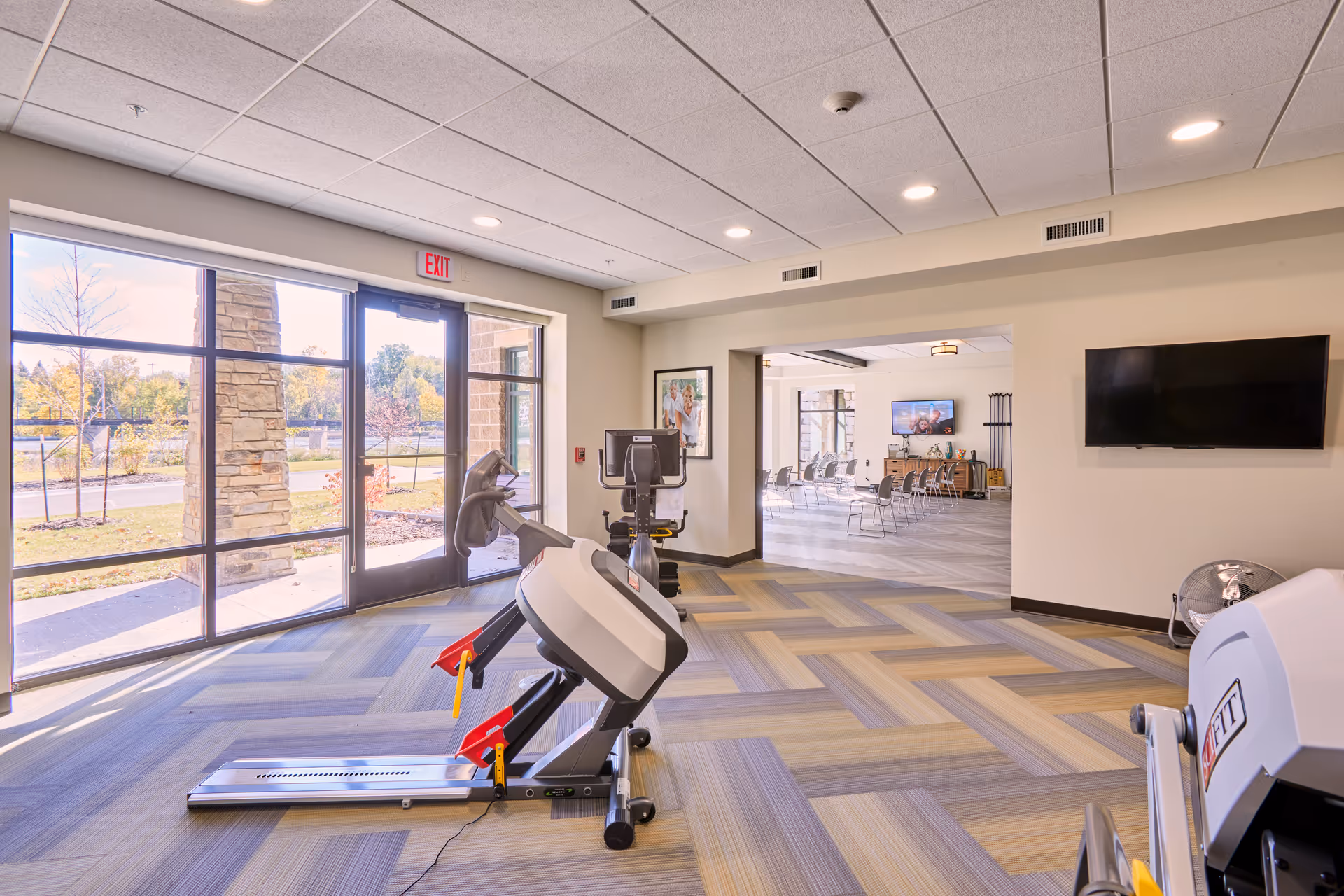 Bright exercise room in a senior living facility with large windows letting in natural light. The room contains exercise equipment including a treadmill and a recumbent bike. Adjacent to the exercise room is a spacious common area with chairs arranged facing a television mounted on the wall.