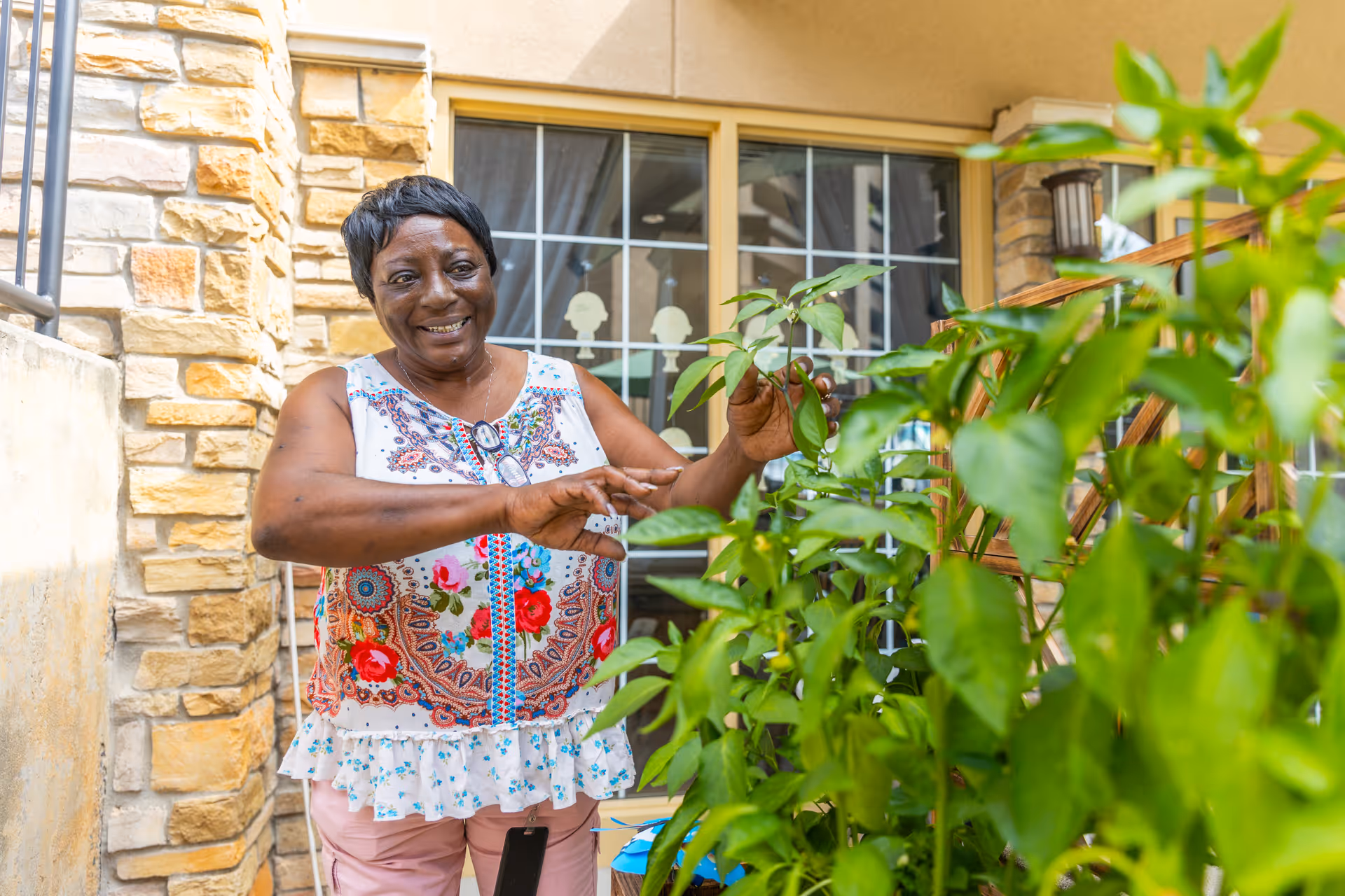 A smiling woman tending green plants on a patio in front of a stone wall and window.