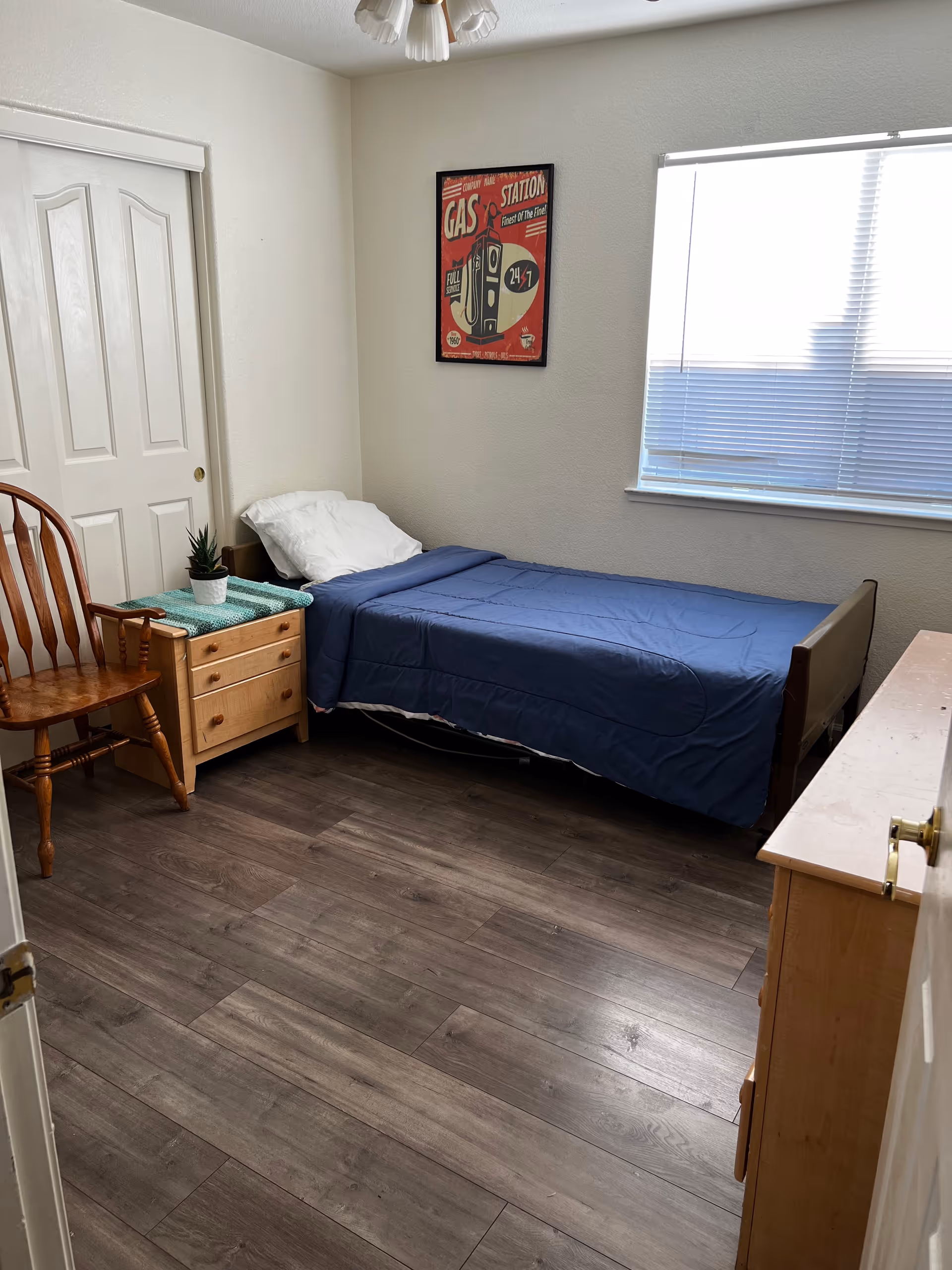 A small bedroom with a single bed covered in a blue blanket and a white pillow. Next to the bed is a wooden nightstand with a small potted plant and a green cloth on top. There is a wooden chair beside the nightstand. A framed vintage-style gas station poster hangs on the wall above the bed. A window with closed blinds is on the right wall, and a wooden dresser is partially visible on the right side of the image. The floor is dark wood, and the walls are painted white.