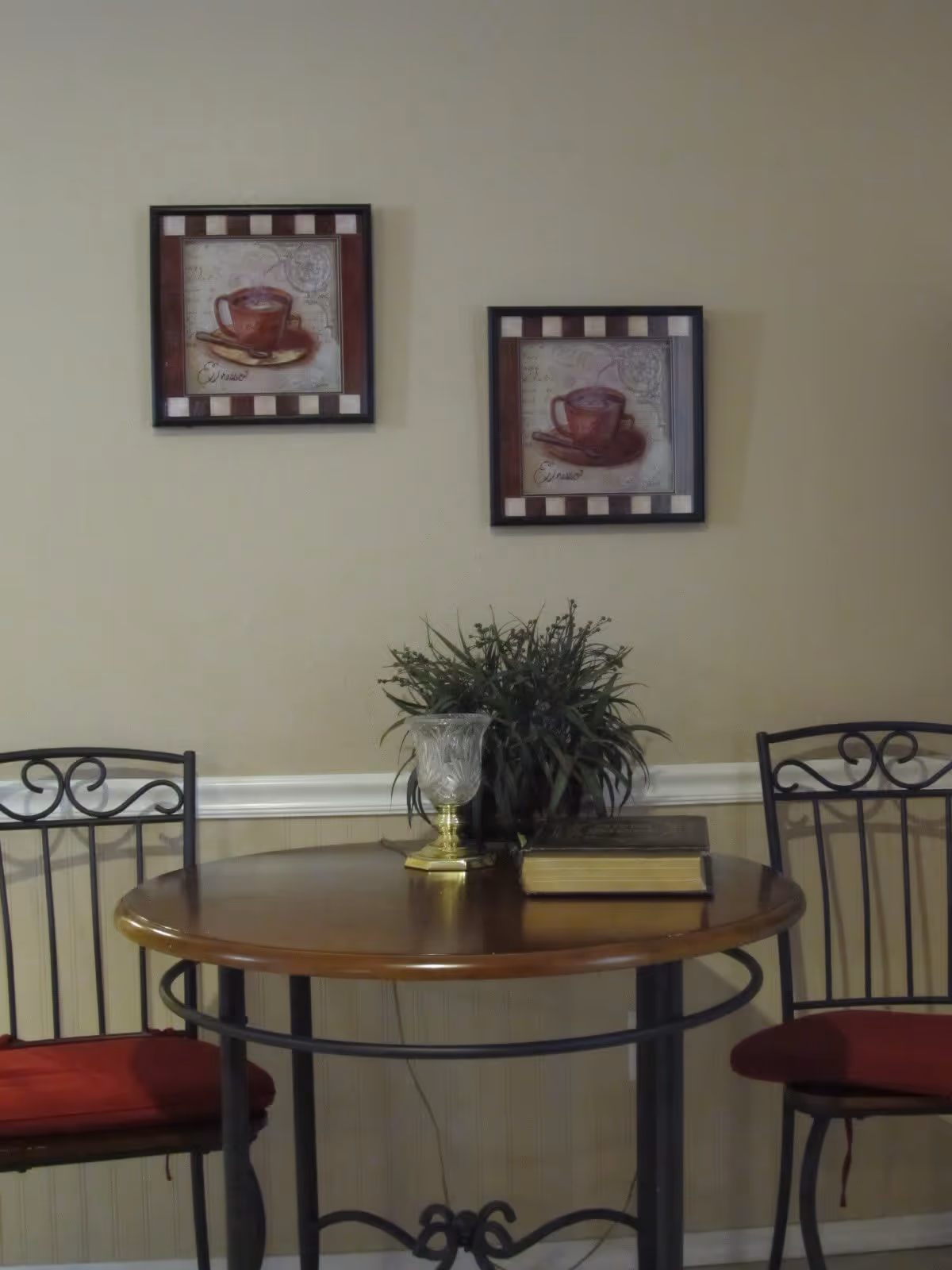 A small round wooden table with a metal frame, flanked by two metal chairs with red cushions. On the table, there is a decorative glass candle holder, a green leafy plant, and a closed book. Two framed pictures of coffee cups hang on the beige wall behind the table.