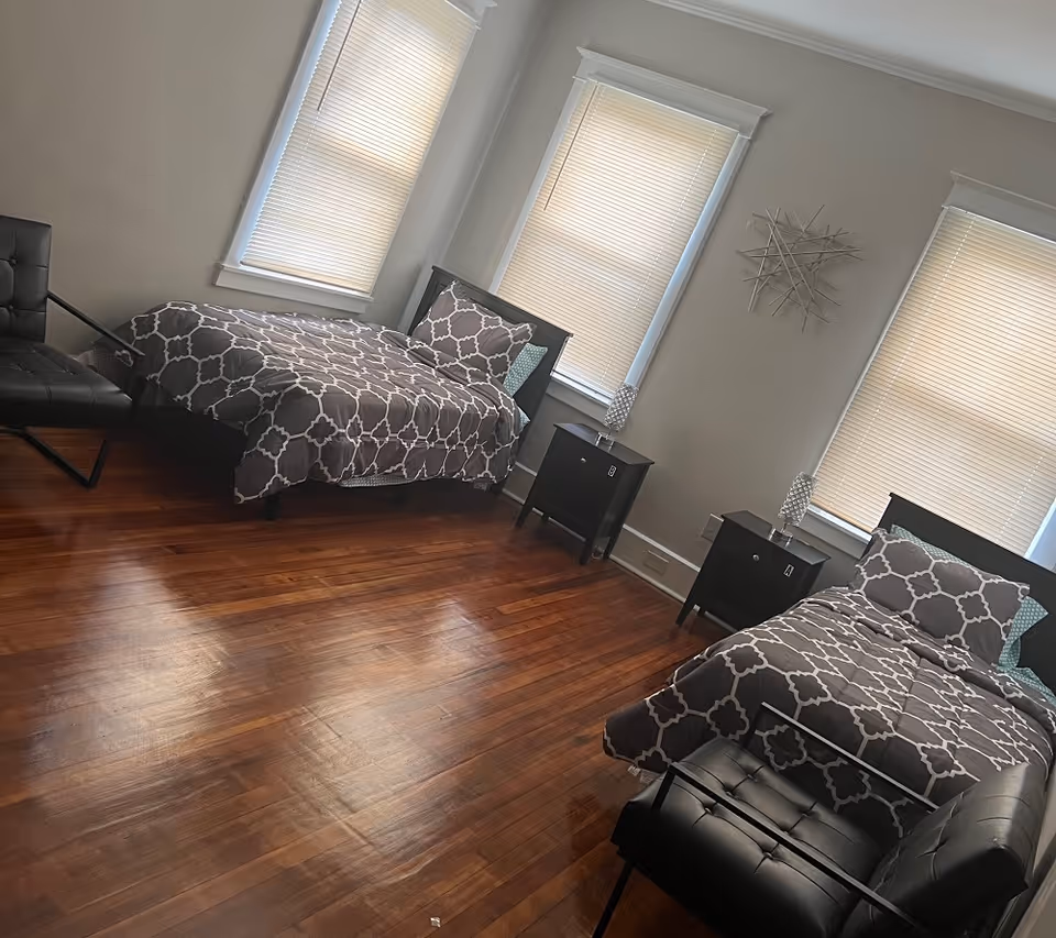 A bedroom with two twin beds, each with a dark wooden frame and matching gray and white patterned bedding. Between the beds are two black nightstands, each with a decorative lamp. The room has three windows with closed blinds and light gray walls. There are two black chairs, one near each bed, and the floor is polished hardwood.