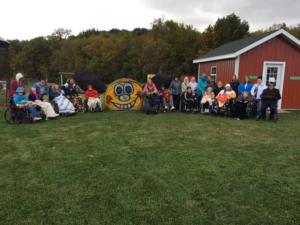 A group of elderly people, some in wheelchairs, and caregivers gathered outside on a grassy area in front of a red building. Behind them is a large painted hay bale with a smiling face and black ears, resembling a cartoon character. Trees with autumn foliage are visible in the background under a cloudy sky.