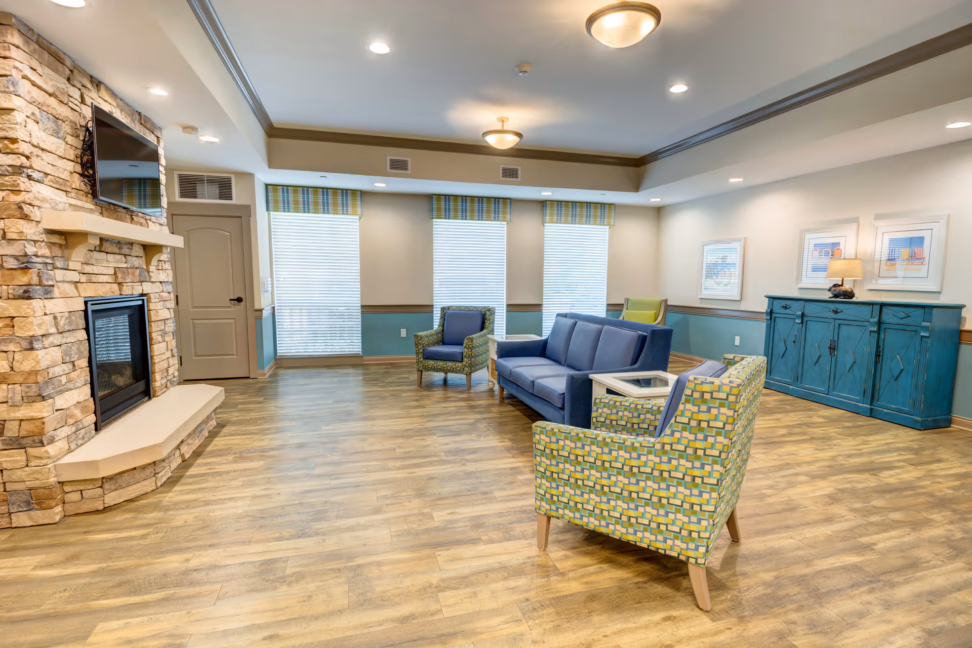 A spacious living room with wood flooring, a stone fireplace with a mounted TV above it, and several chairs and a sofa arranged around a small table. The room has three large windows with blinds and striped valances, a blue sideboard with a lamp and framed artwork above it, and recessed ceiling lights.