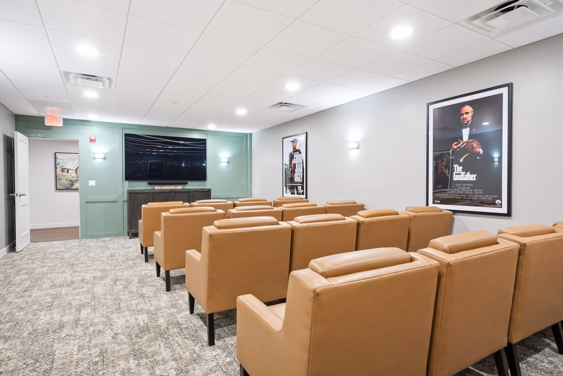 A small theater room with rows of tan leather chairs facing a large flat-screen TV mounted on a green accent wall. The room has light gray walls with framed movie posters, including The Godfather, and soft wall sconces providing lighting. The floor is carpeted in a patterned design, and there is an open door leading to another room.