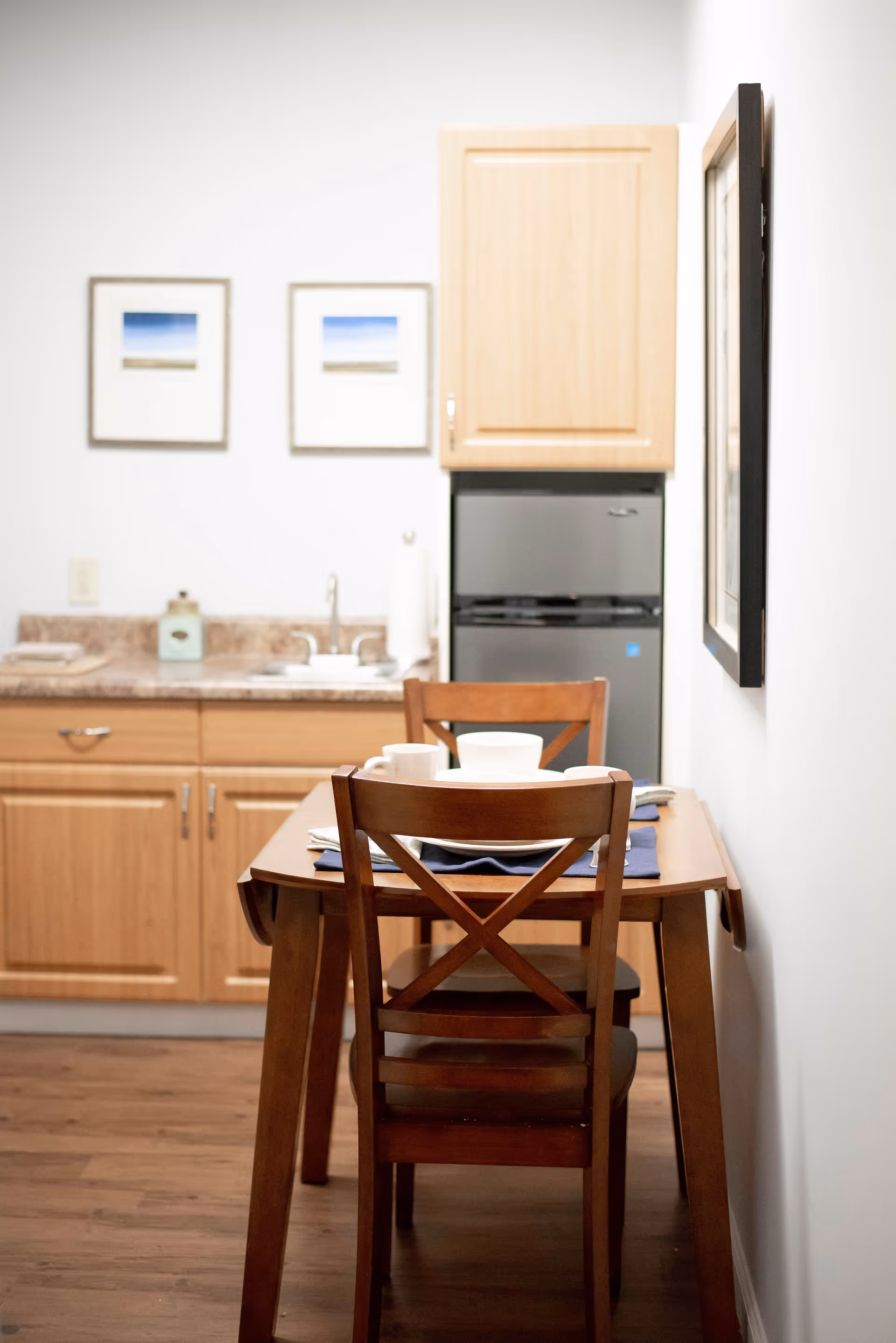 Small kitchen area with wooden cabinets, a countertop with a sink, a mini refrigerator, and a wooden dining table with two chairs. The table is set with white dishes and blue napkins. Two framed pictures hang on the wall above the countertop.
