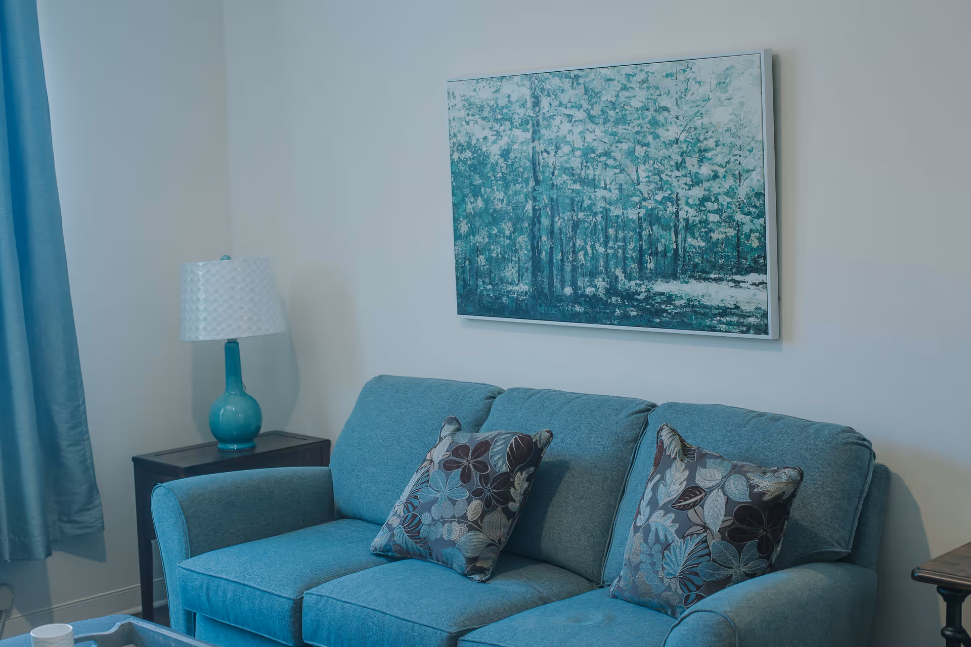 Teal sofa with patterned pillows beneath a framed forest painting, next to a side table and lamp in a living room.