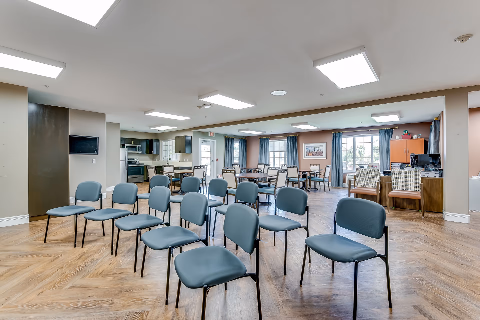 A spacious, well-lit common area in a senior living facility featuring rows of blue chairs arranged in the foreground, several tables and chairs in the background near large windows with blue curtains, and a kitchen area with appliances and cabinetry on the left side.