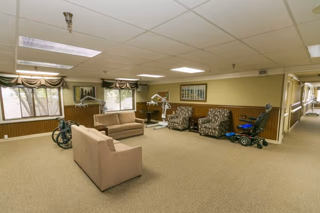 Spacious common sitting area with sofas, armchairs, mobility aids and large windows in a senior living facility.