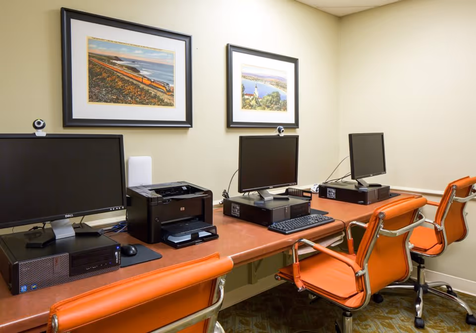 A small computer room with three desktop computers and a printer on a long brown desk. Each computer has a monitor, keyboard, and mouse, with orange office chairs in front of them. Two framed landscape paintings hang on the beige wall above the desk.