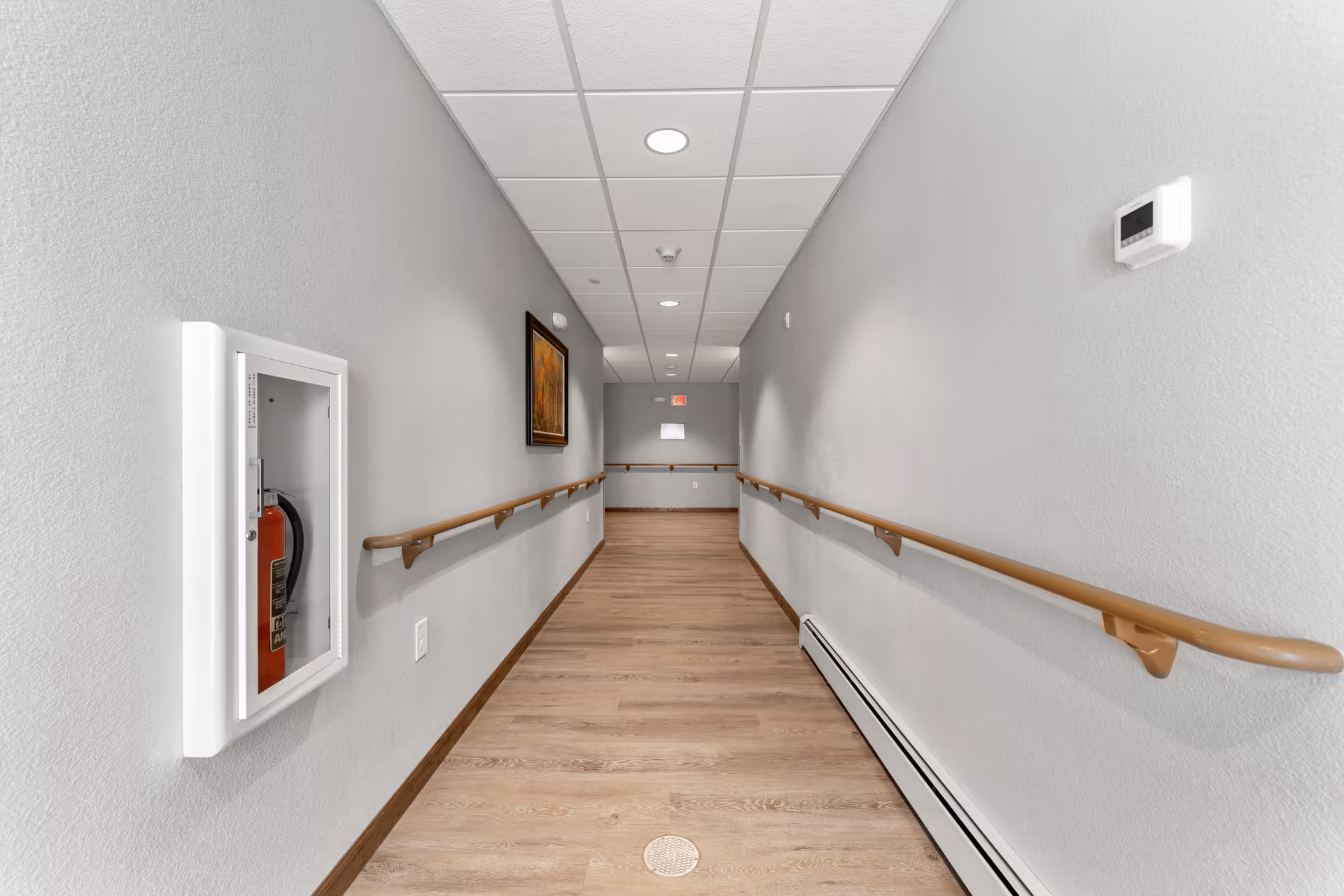A long, narrow hallway with light gray walls, wooden handrails on both sides, a wooden floor, and a white ceiling with recessed lighting. A fire extinguisher is mounted on the left wall inside a white case, and a framed picture hangs on the left wall. A thermostat is mounted on the right wall.
