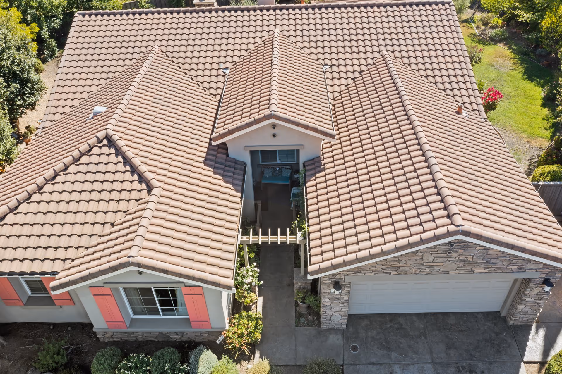 Aerial view of a single-story house with a tiled roof, a small front porch with seating, a driveway leading to a two-car garage, and surrounding greenery and garden areas.
