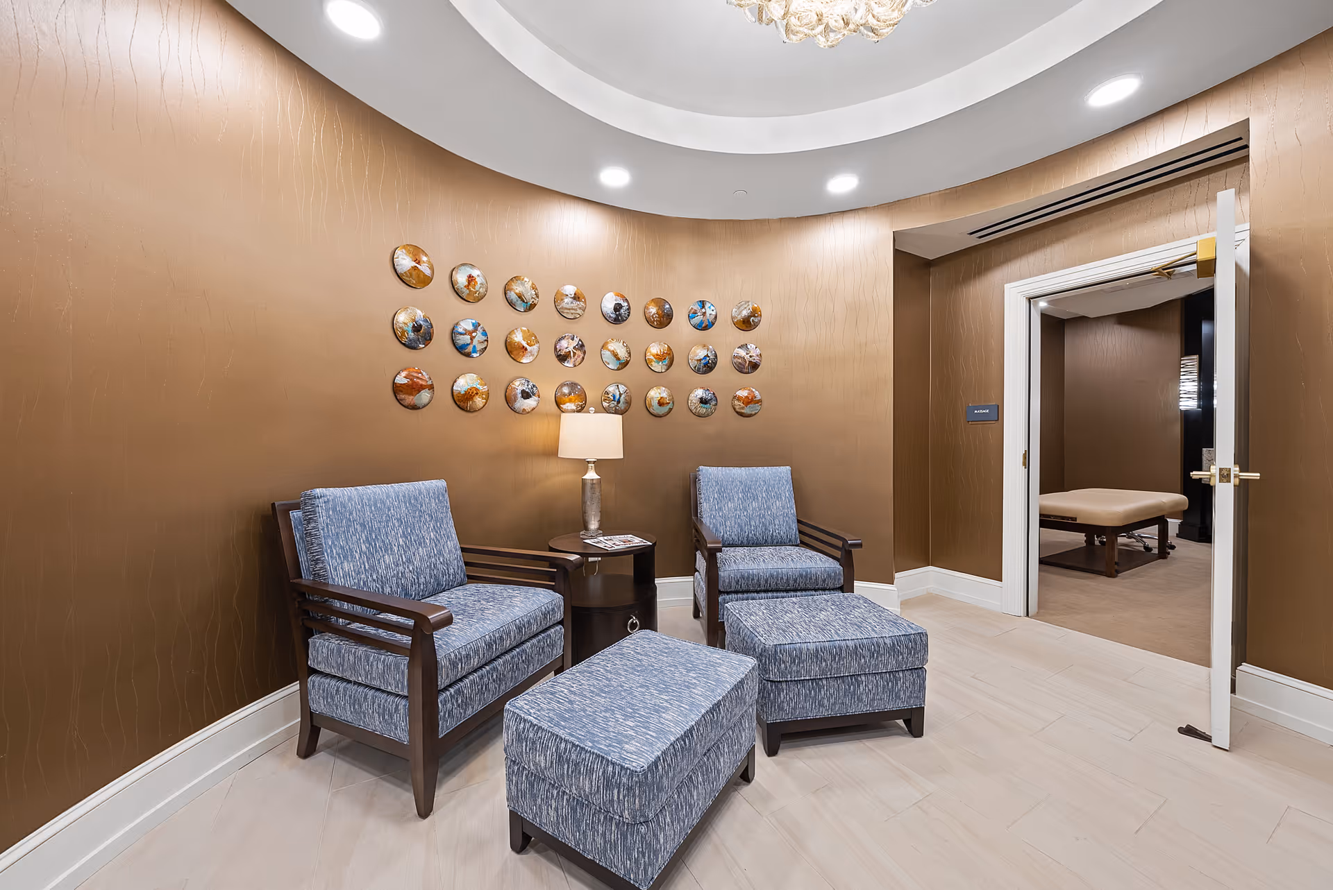 A cozy seating area with two blue upholstered armchairs and matching ottomans arranged around a small round wooden table with a lamp. The walls are painted brown with a decorative circular art installation above the table. An open door leads to another room with a massage table visible inside.