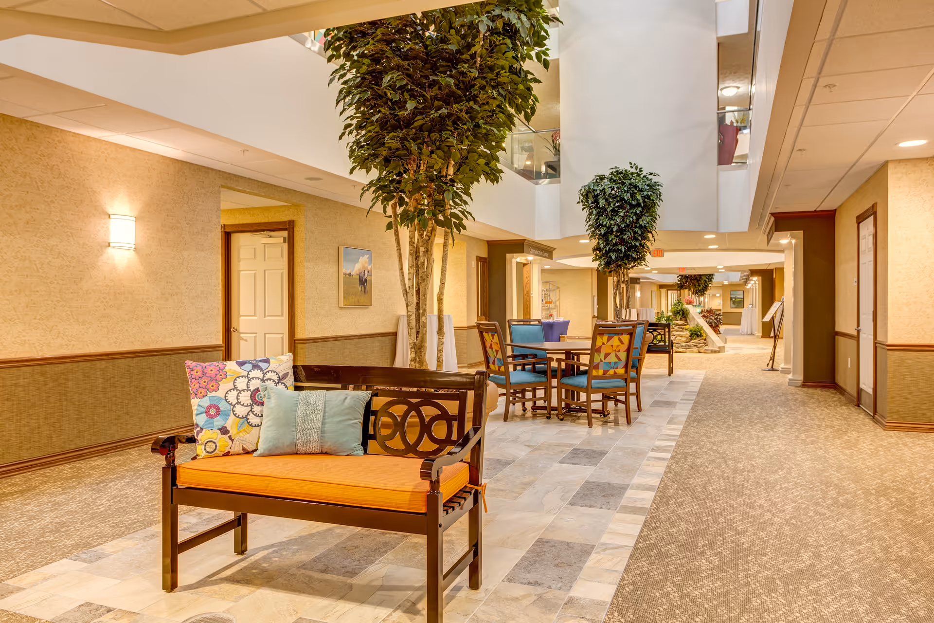 A well-lit indoor common area with a wooden bench featuring colorful cushions in the foreground. There are tall potted plants and a round table with four chairs further back. The floor is a combination of carpet and tiled sections, and the walls are decorated with light-colored wallpaper and framed pictures. The space appears spacious and inviting with a high ceiling and multiple doorways.
