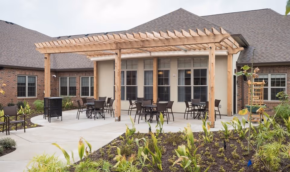 Outdoor patio area at The Pearl at Jamestown - Memory Care Community featuring a wooden pergola with several tables and chairs underneath. The patio is surrounded by landscaped garden beds with various plants and shrubs, and the building's brick exterior and windows are visible in the background.