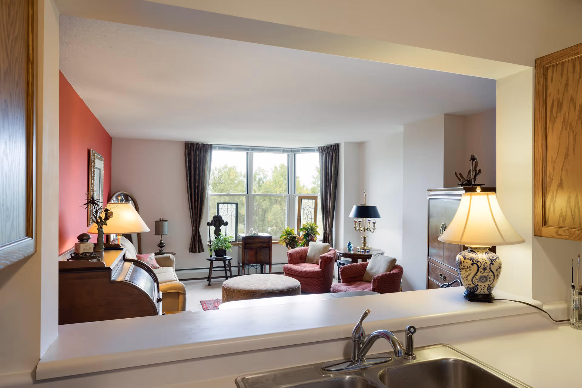 View from a kitchen pass-through window into a cozy living room with a large window, two red armchairs, a round ottoman, a beige sofa, various lamps, and decorative plants.