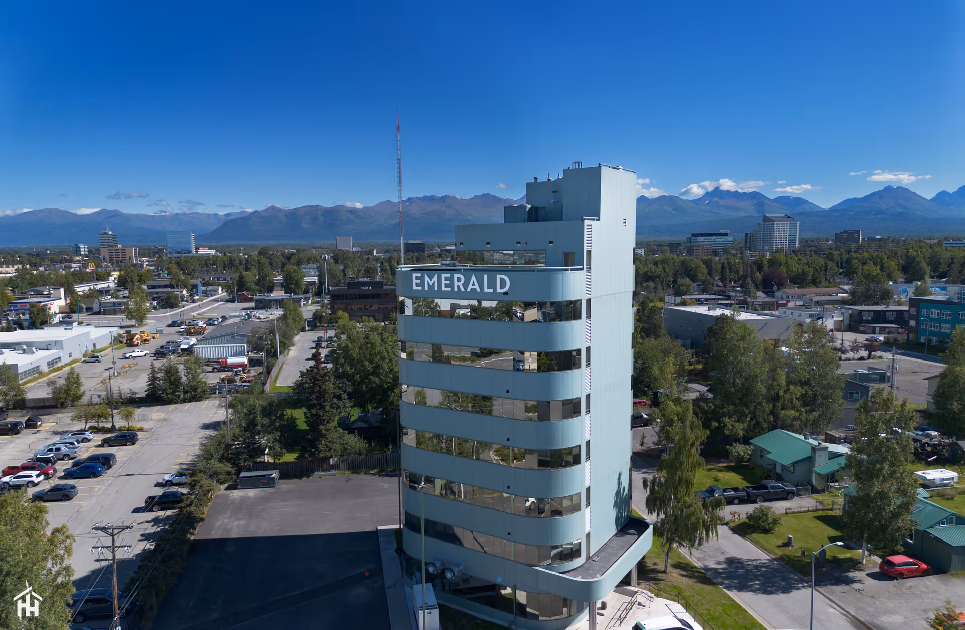 A tall, modern building with reflective windows and the word 'EMERALD' displayed near the top, surrounded by trees, parking lots, and other buildings with mountains visible in the background under a clear blue sky.