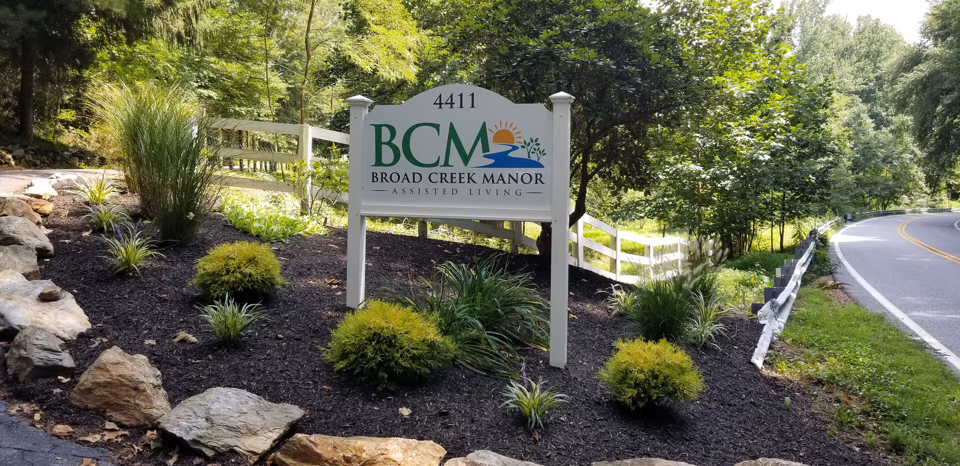 A landscaped area beside a road featuring a white sign with green and blue text that reads '4411 BCM Broad Creek Manor Assisted Living' with a sun and water graphic. The area has small bushes, plants, rocks, and a white wooden fence, surrounded by trees and greenery.