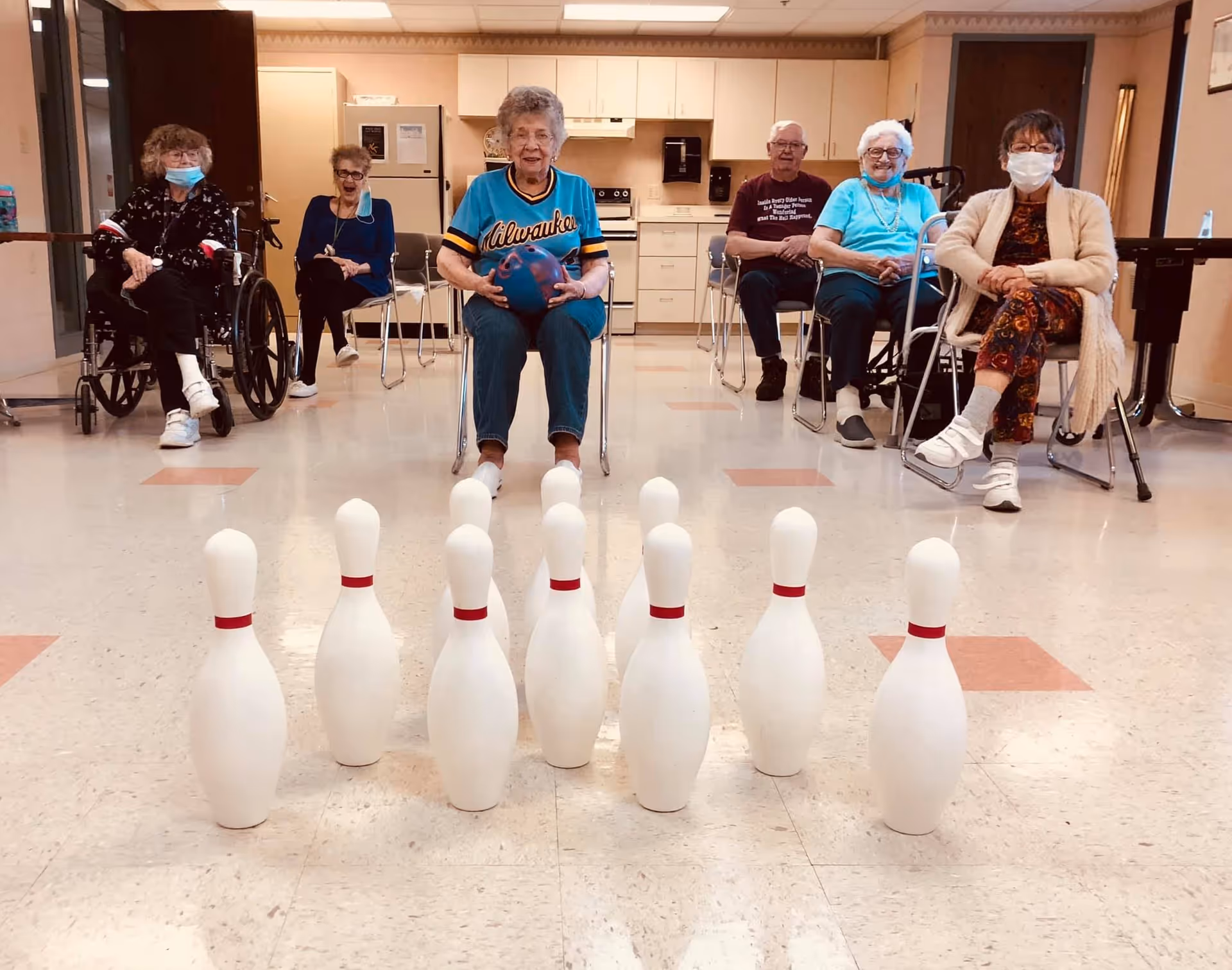 A group of six elderly people sitting in chairs in a room with a kitchen area in the background. One woman in the center is holding a bowling ball, preparing to bowl at a set of ten white bowling pins arranged on the floor. Two of the individuals are wearing face masks.