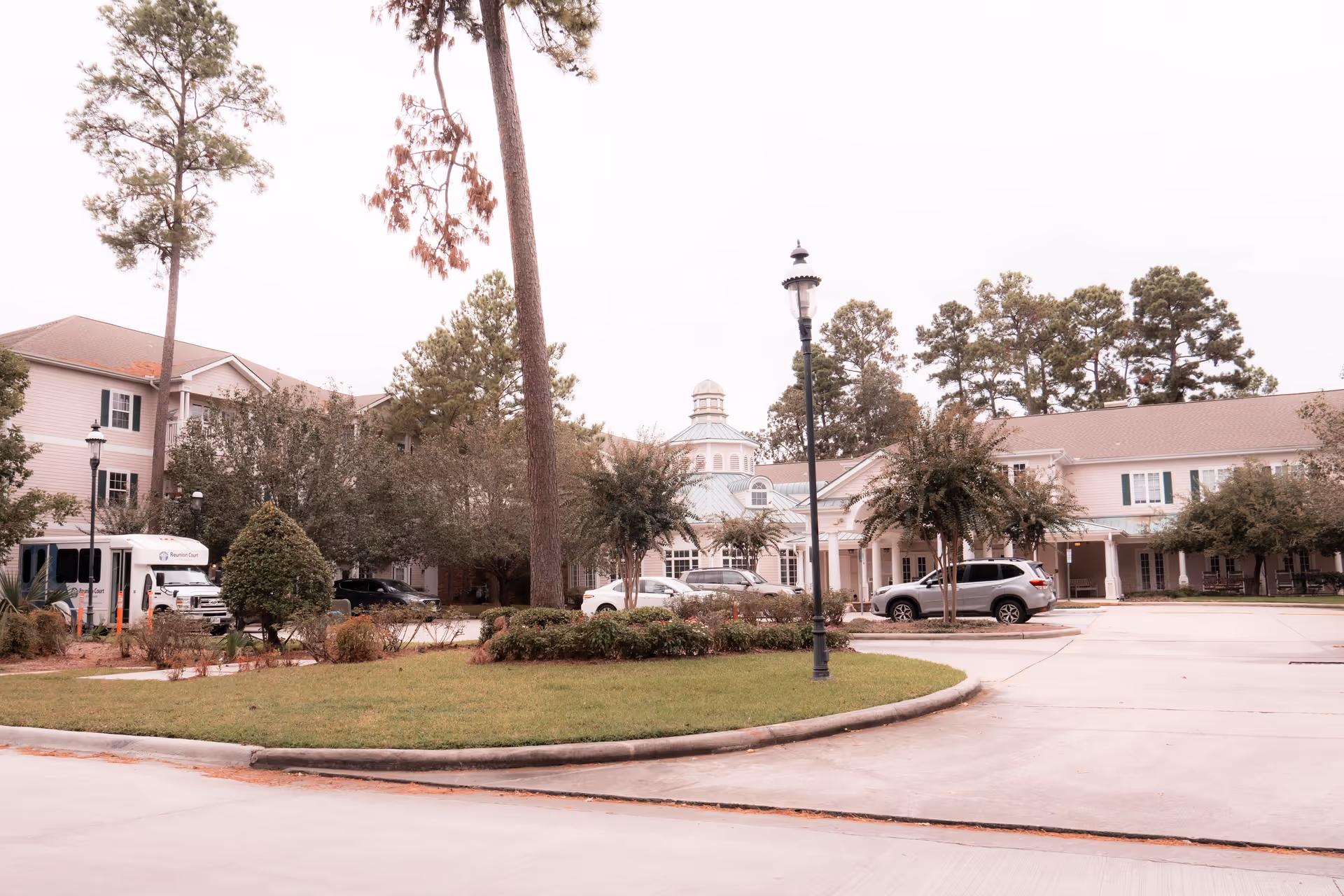 Exterior view of a senior living facility with a circular driveway, landscaped greenery, tall trees, and several parked cars. The building is two stories with a light-colored facade and a central cupola on the roof.