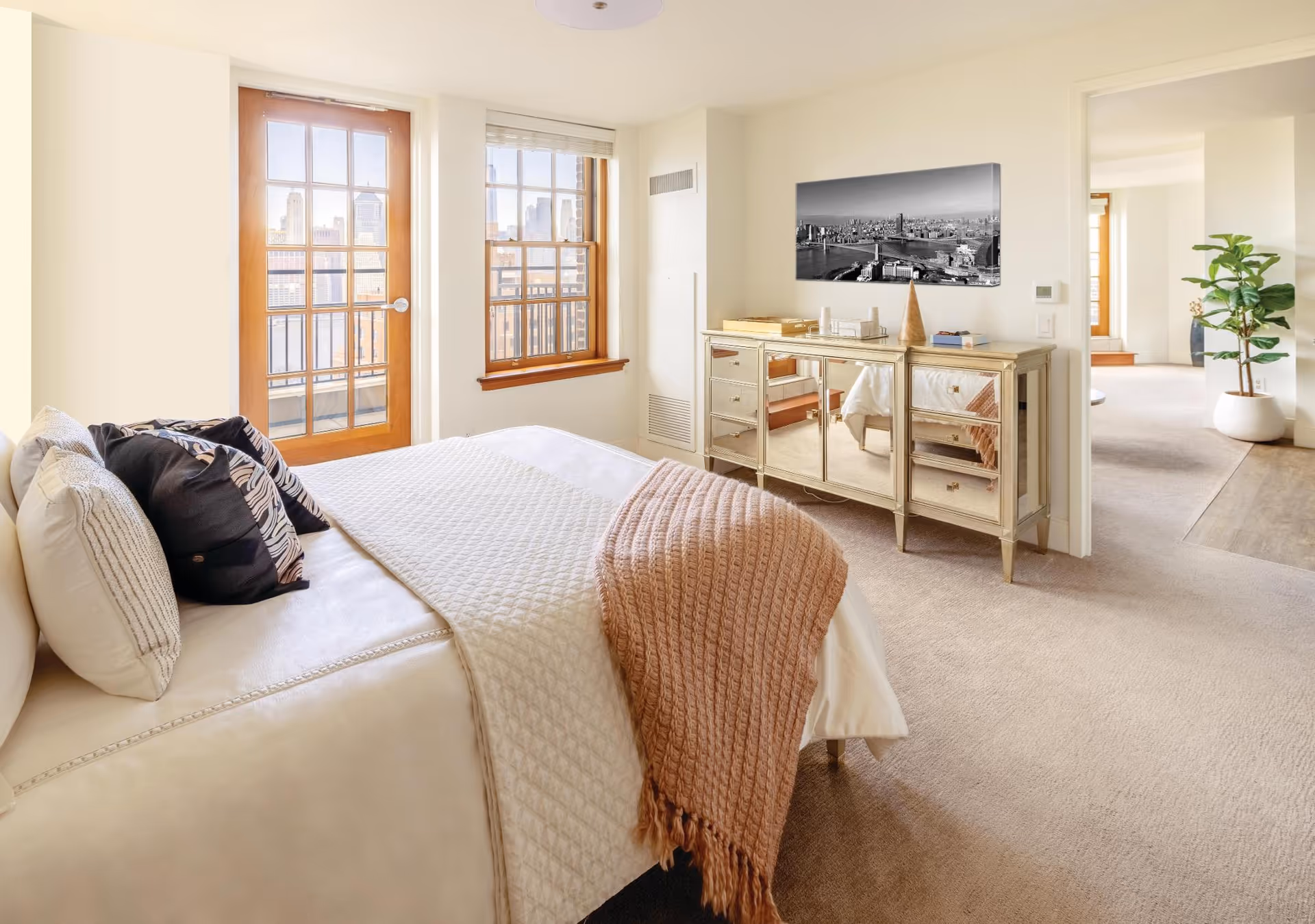 Sunlit bedroom with a neatly made bed, wooden French doors and windows, a mirrored dresser, and an open doorway to an adjacent sitting area.