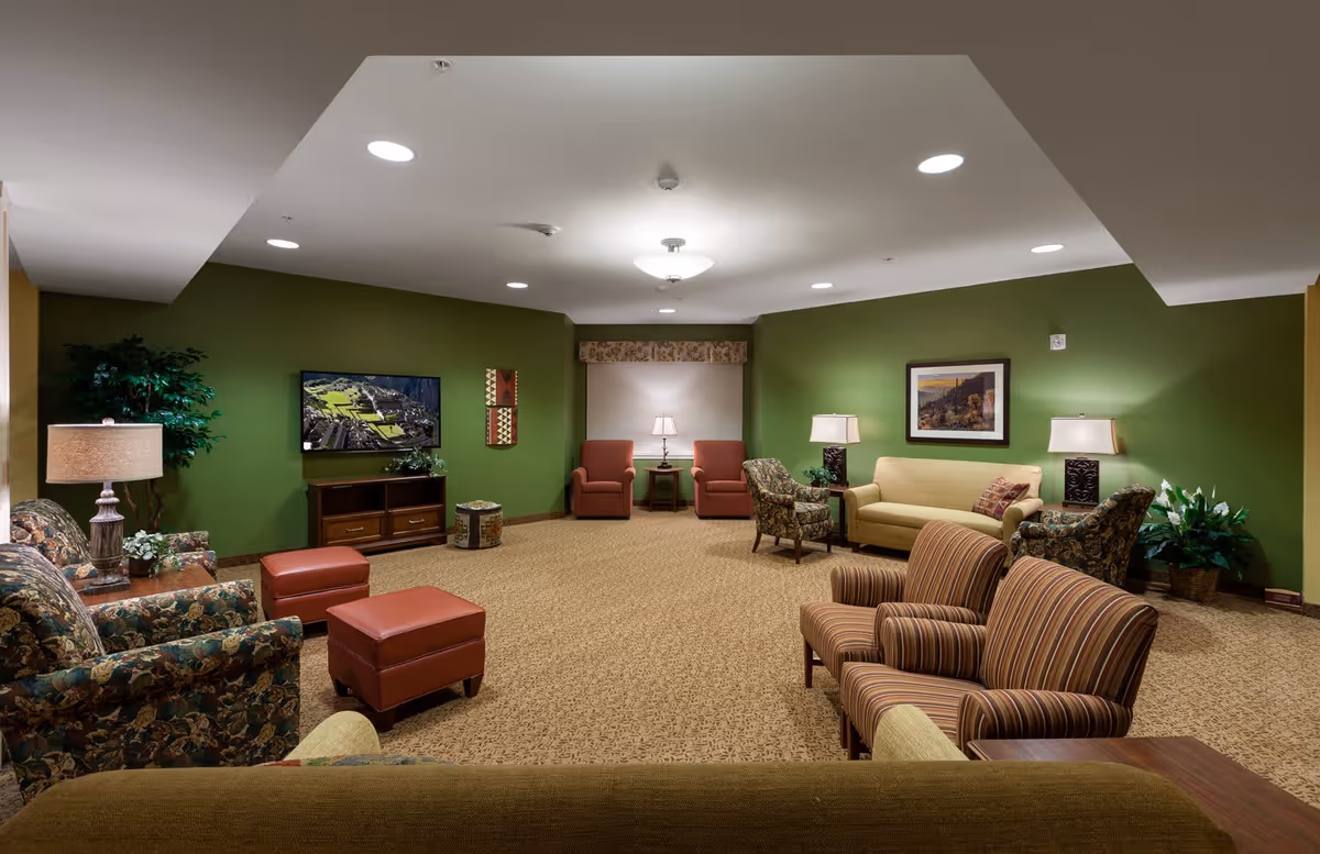 A spacious living room with green walls and beige carpet. The room is furnished with various patterned and solid-colored armchairs, a beige sofa, two red armchairs, and matching red ottomans. There are several table lamps and a flat-screen TV mounted on the wall. Decorative plants and framed artwork add to the cozy atmosphere.