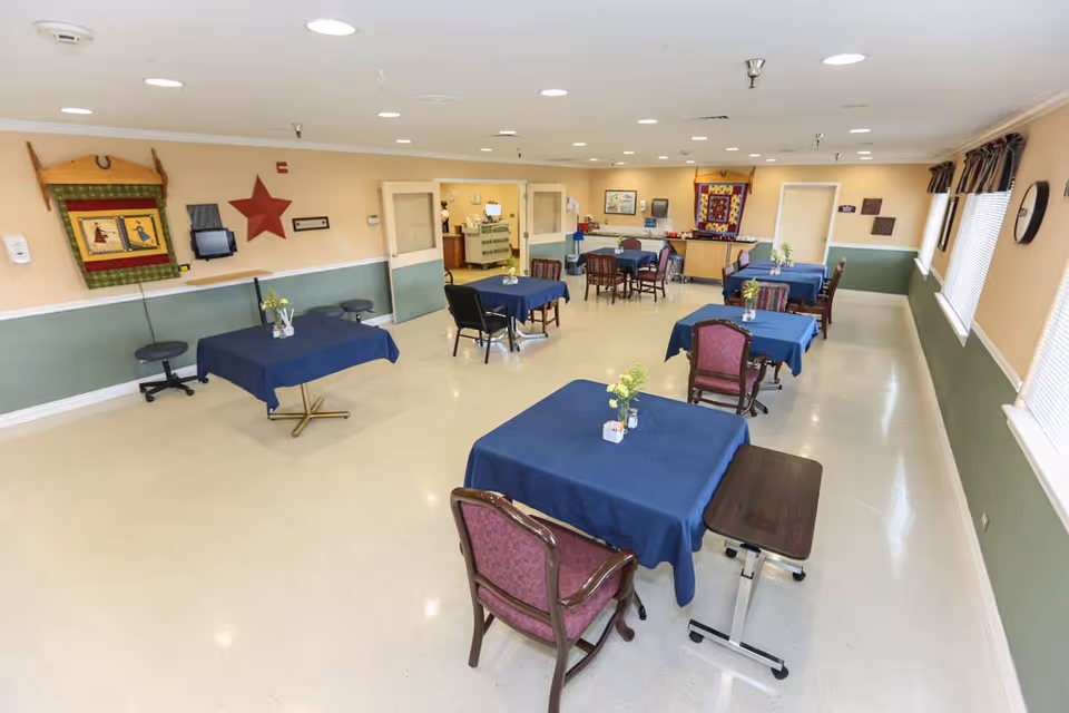 A spacious dining room with several tables covered in blue tablecloths, each adorned with small flower arrangements. The room has beige walls with a green lower half, several windows with blinds on the right side, and various decorative items on the walls including a red star and framed artwork. Chairs with different upholstery styles are arranged around the tables. The room is well-lit with ceiling lights and has a clean, polished floor.