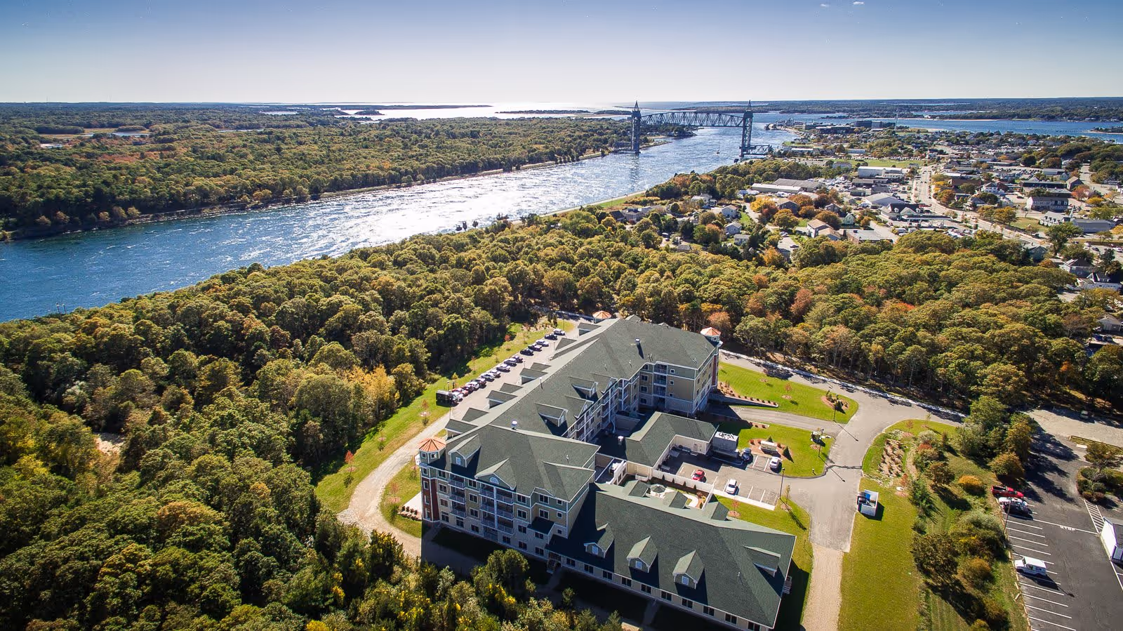 Aerial view of Keystone Place at Buzzards Bay, a large multi-story building surrounded by trees and greenery, with a river and a bridge in the background under a clear sky.