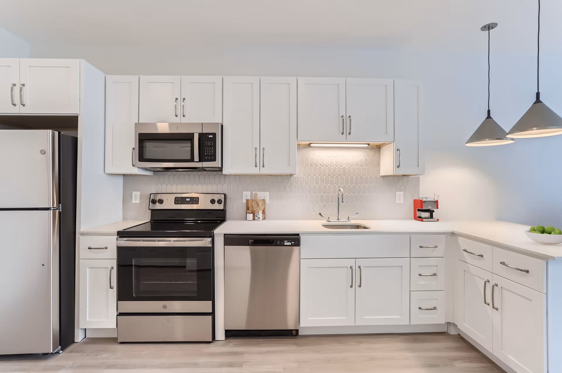 Modern kitchen with white cabinets, stainless steel appliances including a refrigerator, microwave, stove, and dishwasher. The backsplash features a light gray geometric tile pattern. There is a sink under a cabinet with under-cabinet lighting, two pendant lights hanging from the ceiling, a red coffee maker on the counter, and a bowl of green apples on the right side of the counter.