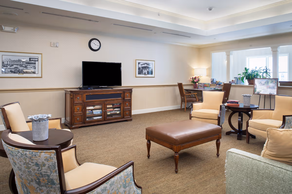 A cozy living room area with beige and patterned armchairs arranged around a brown leather ottoman. A wooden TV stand with a flat-screen television is against the wall, flanked by framed black and white photographs. A clock is mounted above the TV. In the background, there is a desk with a chair, a lamp, and some plants near a window.