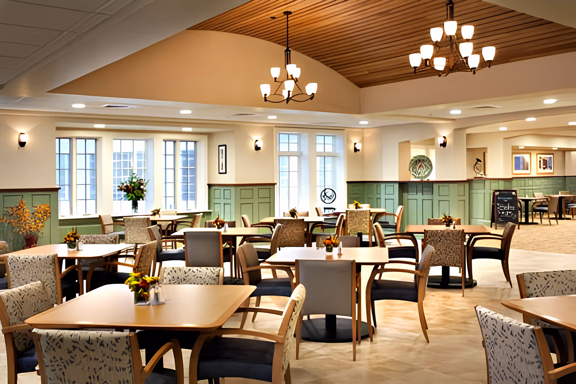 A spacious and well-lit dining room with multiple wooden tables and cushioned chairs arranged neatly. The room features large windows allowing natural light to enter, green wainscoting on the walls, and decorative chandeliers hanging from a wooden ceiling. Small flower arrangements are placed on each table, creating a welcoming atmosphere.