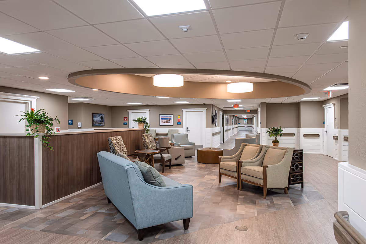A spacious and well-lit common area in a senior living facility featuring comfortable seating including a blue sofa, beige armchairs, and patterned chairs arranged around small tables. The area has a modern ceiling light fixture, plants on counters and tables, and a long hallway with doors and handrails extending into the background.