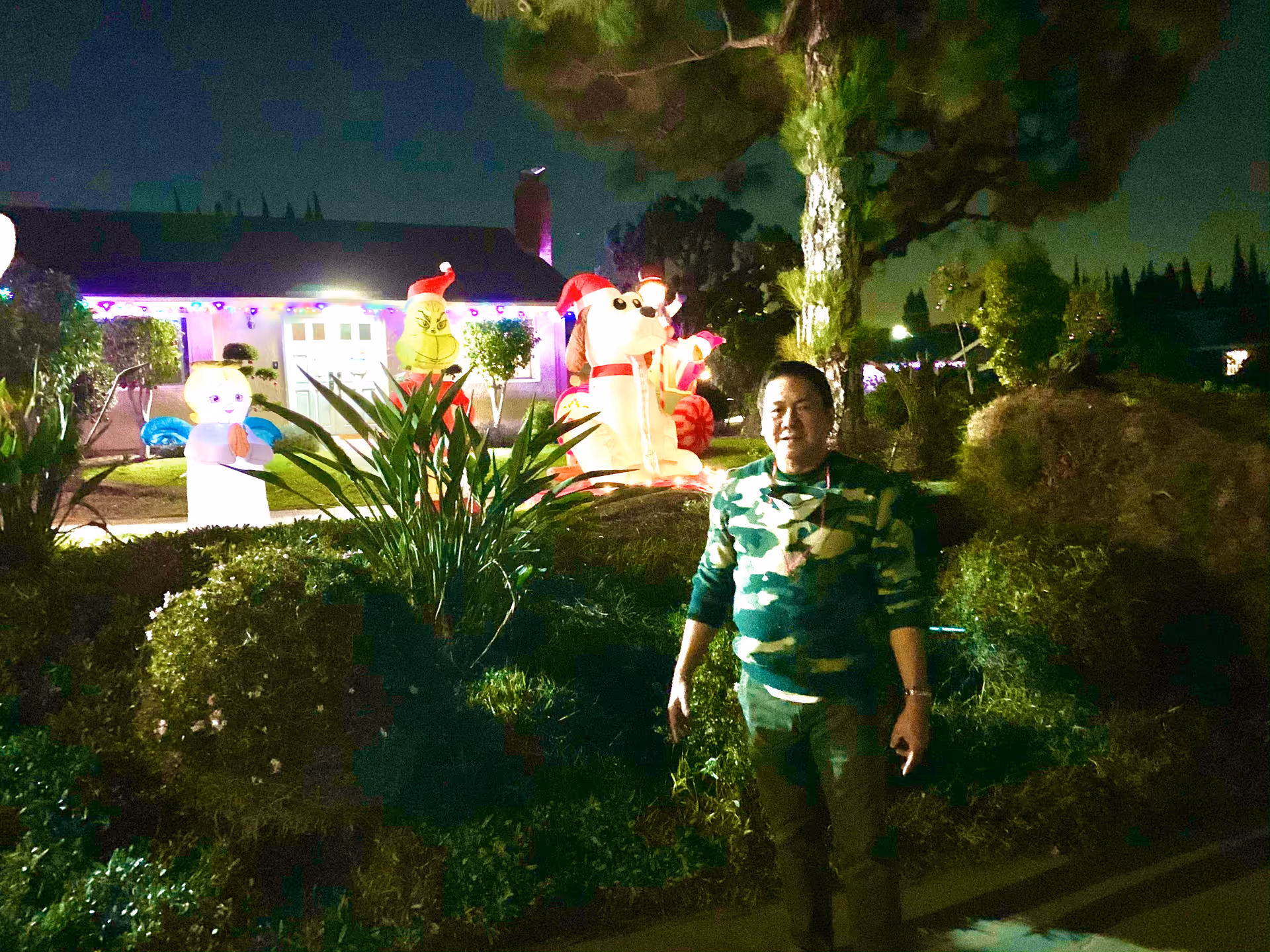 A man wearing a camouflage sweater stands outside at night in front of a house decorated with illuminated holiday inflatables including an angel, the Grinch, and a dog wearing a Santa hat. The yard is landscaped with bushes and trees.