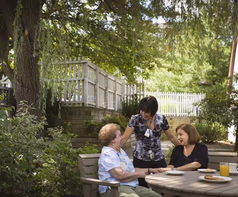 Two elderly women sitting at a round outdoor table with plates of food and a glass of orange juice, while a caregiver stands beside them smiling and engaging in conversation. The setting is a shaded garden area with trees, bushes, and a white picket fence in the background.