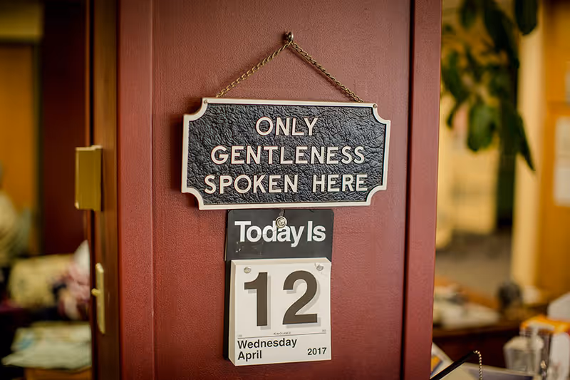 A wall calendar showing the date Wednesday, April 12, 2017, hanging on a reddish-brown wall. Above the calendar is a decorative sign that reads 'ONLY GENTLENESS SPOKEN HERE'. The background shows a blurred indoor setting with furniture and plants.