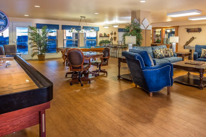 Communal lounge in a senior living facility with blue sofas, a card table, pool table, and shuffleboard on wood floors.