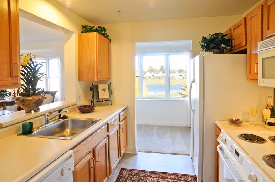 Bright kitchen with wooden cabinets, a sink and white appliances facing a window with a pond view.