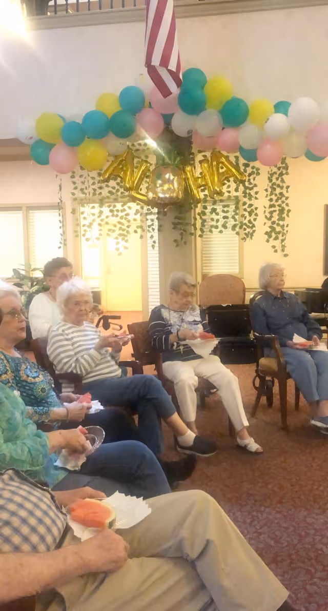 A group of elderly people sitting in a circle in a decorated room with colorful balloons and gold letter balloons spelling 'ALOHA'. They are holding plates with slices of watermelon. The room has hanging greenery and an American flag above.