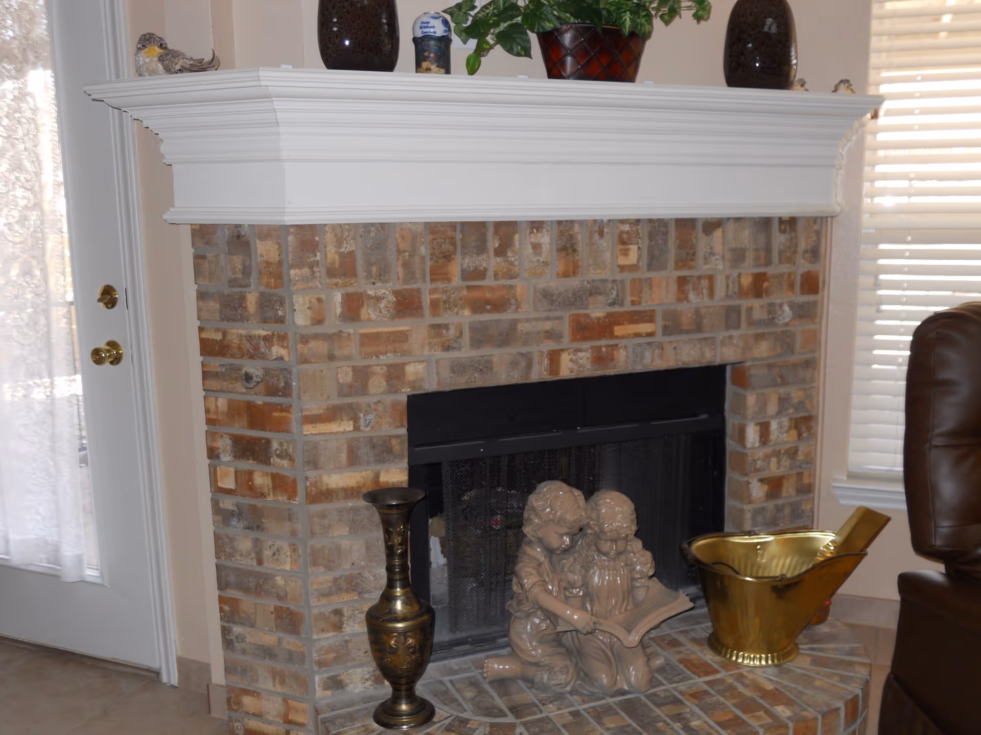 Brick fireplace with a white mantel and a decorative figurine of two children reading, flanked by brass accessories in a living room.