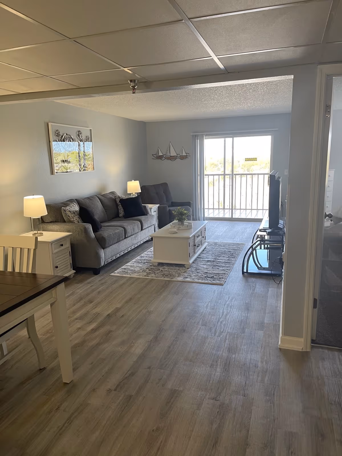 A modern living room with light wood flooring, a gray sofa, a matching armchair, two white side tables with lamps, a white coffee table on a patterned rug, a flat-screen TV on a glass stand, and a sliding glass door leading to a balcony. There is a dining table with chairs partially visible on the left and a bedroom entrance on the right.